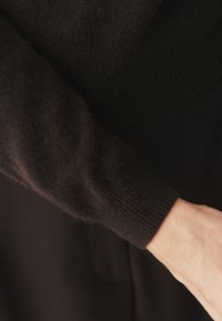 Fekete kötött pulóver ujja ribbelt mandzsettával, puha tapintású és diszkrét fényű. Közeli felvétel, amely kiemeli az anyagot és a dizájn részleteit.