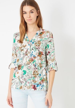 Blusa floral en tejido ligero, con una tapeta de botones, mangas tres cuartos y una mezcla de flores verdes, azules y marrones sobre un fondo blanco.