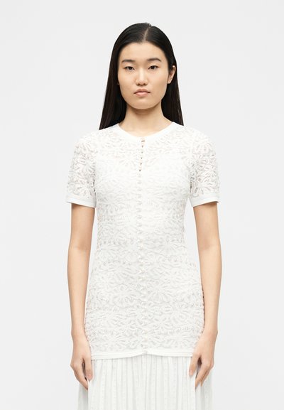 Νεαρή γυναίκα με μακριά σκούρα μαλλιά που φοράει εφαρμοστή λευκή δαντελωτή κοντομάνικη μπλούζα με πετρώματα κουμπιά στο μπροστινό μέρος.