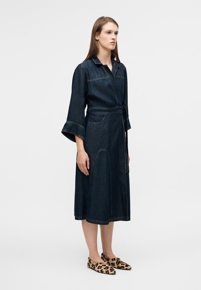 Robe en denim avec manches longues évasées, taille ceinturée et longueur mi-mollet. Tissu bleu foncé avec détails de coutures visibles, associée à des chaussures à imprimé animal.