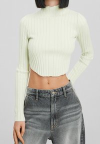 Kvinna iklädd en krämfärgad ribbad långärmad kort tröja och högmidjade blekta svarta jeans, stående med en hand bakom ryggen.