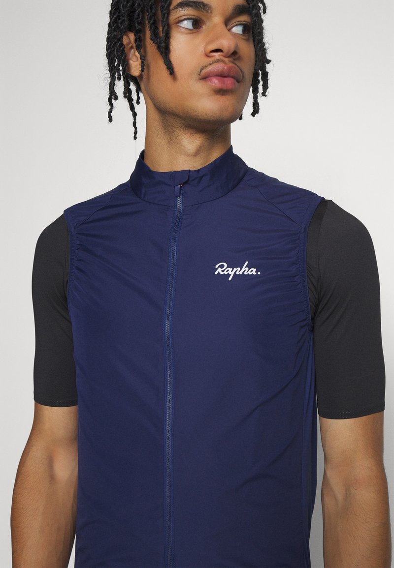 Marine fietsvest met hoge kraag, frontzipper en gefronde textuur op de schouders. Voorzien van het "Rapha" logo in het wit. Zwarte korte mouwen.