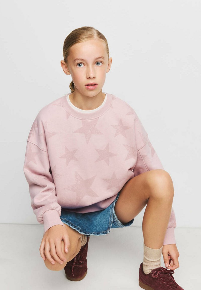 Jeune fille aux cheveux blonds en queue de cheval accroupie, portant un sweat rose à motifs d’étoiles, une jupe en jean, des chaussettes beiges et des chaussures bordeaux.