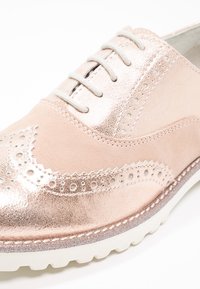 Chaussure en cuir rose avec accents métalliques, détails perforés complexes et tige texturée. Elle présente une semelle en caoutchouc blanche épaisse.