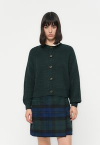 Donkergroene gebreide cardigan met ronde hals en houten knopen, gecombineerd met een geruite rok in groene en blauwe tinten.