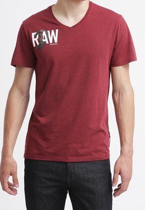 Man met een donkerrood V-hals T-shirt met het tekstlogo "RAW" op de borst, gecombineerd met donkere jeans, getoond van nek tot heupen.