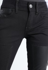 Close-up of black jeans with stitched leather patches on thighs and visible metal button on waistband.