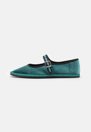 Chaussure Mary Jane en velours turquoise plat avec bordure noire, bride à boucle, détails tressés sur la semelle et semelle extérieure en caoutchouc noir sur fond uni.