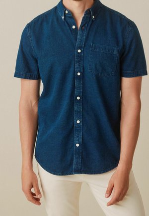 Homme portant une chemise en denim bleu foncé à manches courtes avec une poche poitrine et un pantalon écru sur un fond neutre.
