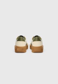 Par et par krem og oliven sneakers med tykke brune gummisåler merket "Sorel," vist fra baksiden på en enkel grå bakgrunn.