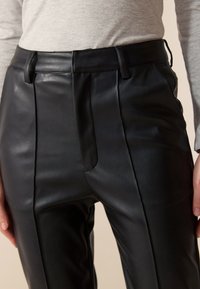 Pantalon en cuir noir avec taille haute, plis, poches latérales et texture lisse, présenté sur un fond neutre.