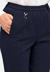 Marineblaue Hose mit strukturiertem Muster, ausgestattet mit einem elastischen Bund und einem silbernen Clipakzent. Auf der rechten Seite ist eine Taschen sichtbar.