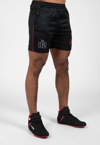Shorts de sport noirs avec des rayures latérales rouges et "Est. 82" imprimé en blanc. Associés à des chaussures noires avec des accents rouges et une sangle.