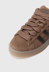 Brown suede sneaker with thick, brown laces, rounded toe, three dark stripes on the side, and a rubber sole with a slight translucent finish.