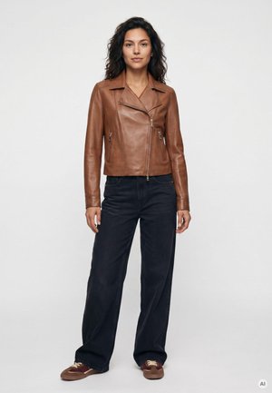 Frau trägt eine braune Lederjacke, dunkle weit geschnittene Jeans und braune Sneakers, steht vor einem schlichten weißen Hintergrund.
