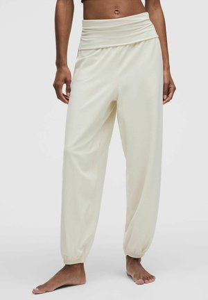 Persona che indossa pantaloni larghi a vita alta color crema con polsini elastici arricciati, in piedi a piedi nudi su uno sfondo semplice.