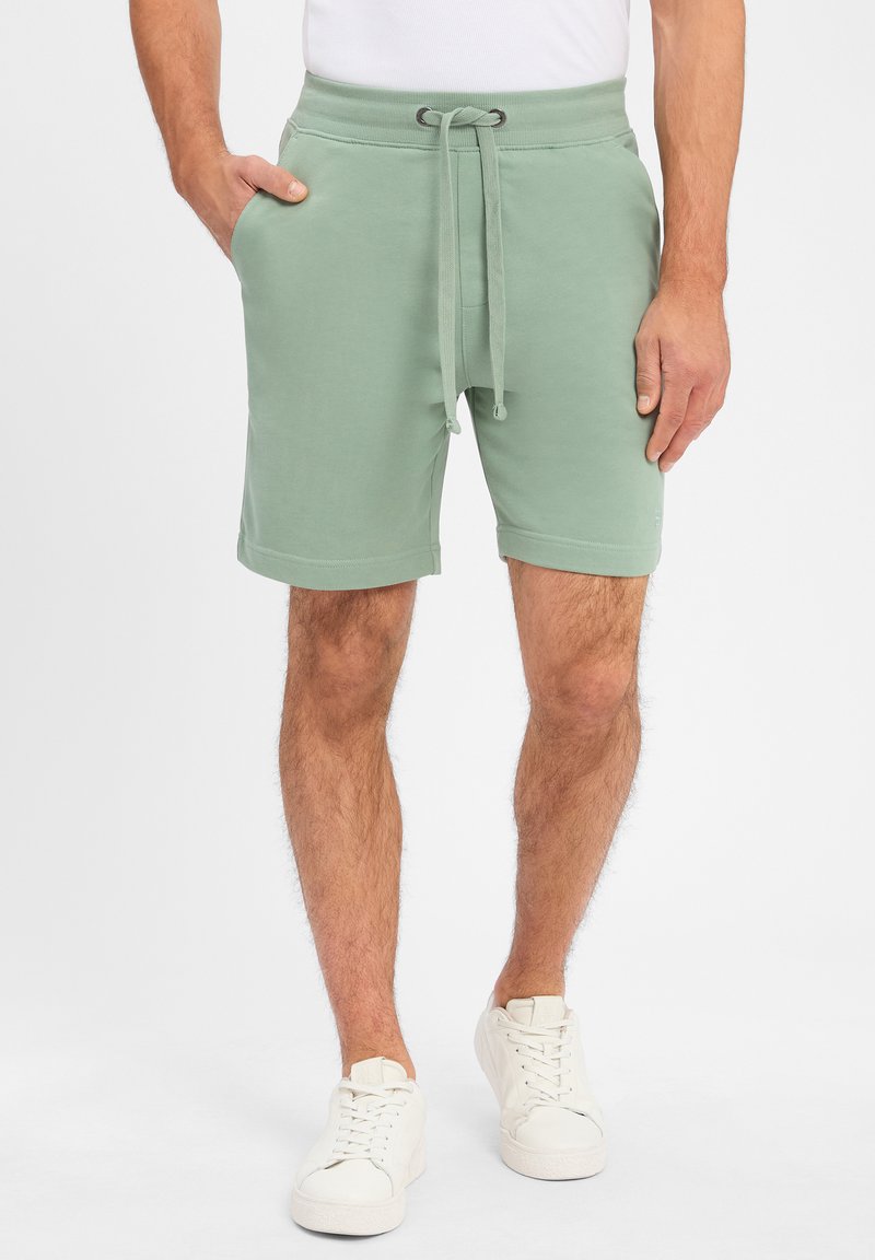 Grüne Baumwollmischung-Shorts mit elastischem Bund, Kordelzugverschluss, seitlichen Taschen und aufgeschlagenen Säumen. Weiße Turnschuhe runden den Look ab.