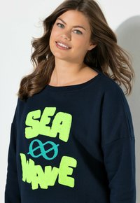 Junge Frau mit welligem braunem Haar lächelt und trägt ein marineblaues Sweatshirt mit leuchtend grünem "SEA WAVE"-Schriftzug und einem blauen Knotenmuster auf der Vorderseite.
