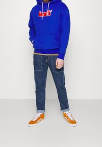 Sudadera azul con el logo rojo de "Levi's", combinada con jeans de mezclilla oscura y zapatillas de ante naranjas. La sudadera cuenta con un bolsillo frontal y cordones.