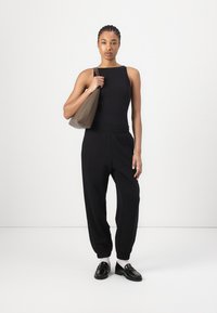 Black ribbed tank top and black jogger pants, paired with black loafers. The model holds a soft, beige shoulder bag. Clean, minimalistic design.