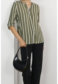 Personne portant une chemise rayée olive et blanche et un pantalon noir, tenant un sac à main noir en forme de demi-lune par sa poignée.