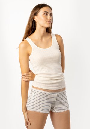 Junge Frau mit langem braunem Haar trägt ein weißes ärmelloses Top und gestreifte Shorts, steht mit einem leicht angewinkelten Arm.