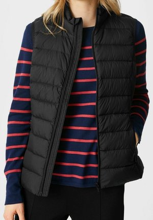 Person iført en sort quiltet vest over en marineblå trøje med horisontale røde striber, hænderne i lommerne, foran en ensfarvet baggrund.