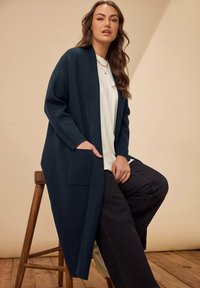 Cardigan lungo blu navy con vestibilità rilassata, dotato di tasche frontali, polsini a coste e una texture morbida, indossato sopra una camicia bianca e jeans scuri.