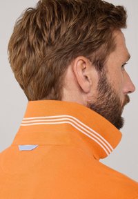 Polo arancione con colletto alto, caratterizzato da strisce bianche. Il materiale sembra testurizzato e morbido, con un piccolo accento blu sul retro.