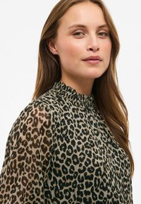 Blusa con stampa leopardata e scollatura a volant, con maniche trasparenti e tessuto leggero. Toni terrosi dominano la palette di colori.