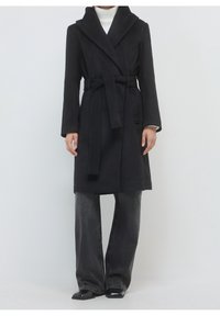 Personne portant un manteau noir ceinturé sur un col roulé blanc, assorti à un jean large foncé et des chaussures noires à bouts pointus, debout sur un fond blanc.