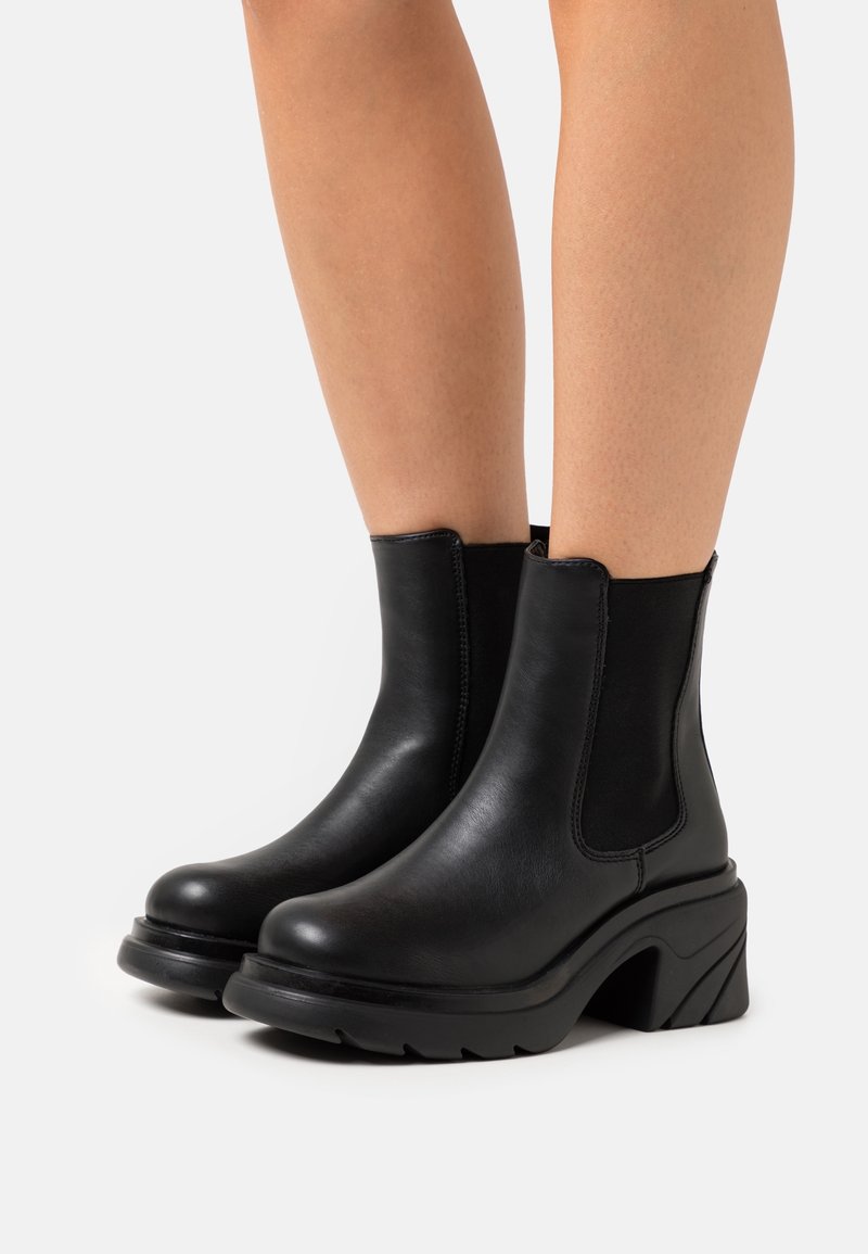 Bottes cheville en cuir noir avec talons épais et panneaux élastiques sur les côtés, portées sur des jambes nues sur un fond blanc.