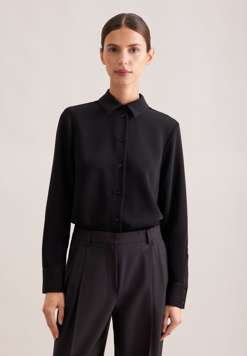 Femme aux cheveux foncés tirés en arrière, portant une chemise noire à manches longues boutonnée et un pantalon sombre taille haute, debout devant un fond uni.