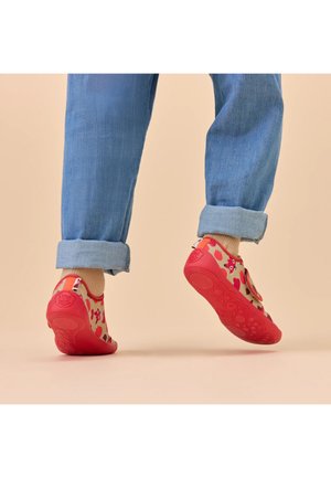 Dziecko noszące beżowo-czerwone buty z wzorami kreskówek i beżowe skarpetki, unoszące stopy nad beżową podłogą, podwinięte niebieskie dżinsy.