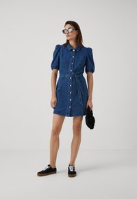 Jeansklänning med puffärmar, knappar framtill, åtstramad midja med ett bälte, längd till mitten av låret. Bärs med svarta sneakers och solglasögon.