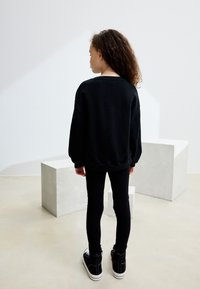 Felpa nera oversize con spalle scese, abbinata a leggings aderenti neri e sneakers nere con dettagli bianchi. Sfondo neutro.