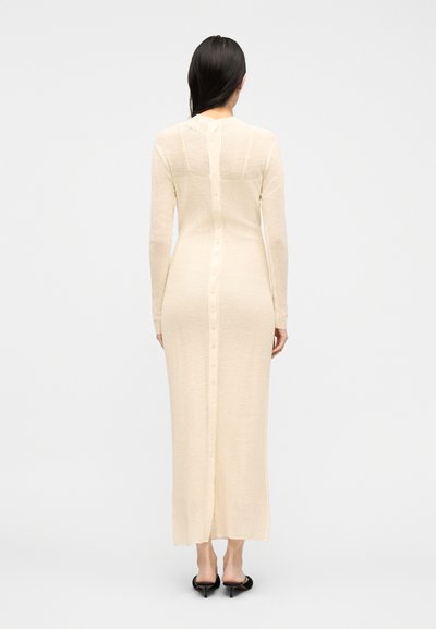 Femme de dos portant une longue robe en maille crème avec des boutons courant le long du dos et des escarpins noirs à bride arrière.