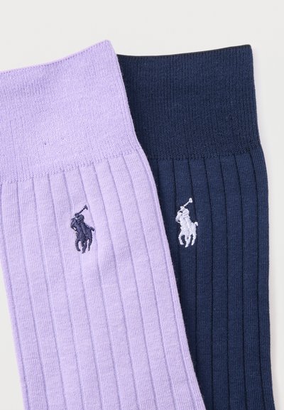 Ζευγάρι ριμπ πλεκτές κάλτσες, μία λεβαντών με σκούρο λογότυπο και μία ναυτικό μπλε με λευκό λογότυπο, διπλωμένες πλάι πλάι σε λευκό φόντο.