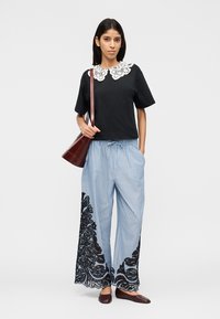 Femeie purtând un top negru cu guler de dantelă albă, pantaloni largi cu dungi albastre și model de dantelă neagră, pantofi maro și o geantă maro purtată pe umăr.