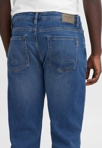 Jeans de mezclilla azul con corte recto, que cuentan con dos bolsillos traseros con costuras en forma de montaña y una etiqueta gris en la cinturilla.
