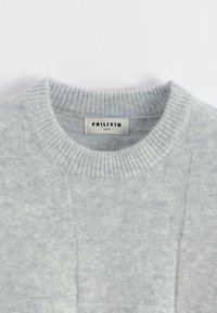 Pull tricoté gris clair avec un col rond côtelé et une étiquette "FRILIVIN" taille M cousue à l'intérieur du col.