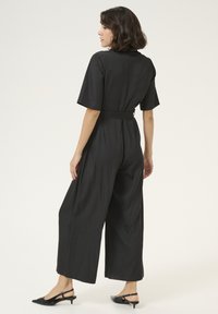 Svart jumpsuit i mjukt tyg, med korta ärmar, vida ben, en figursydd midja med bälte och en ren ryggdesign.