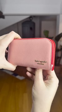 Rožnato roza usnjen denarnica z teksturirano površino, rdečim zadrgo in zlatim logotipom. Drži se v roki za tesen pogled.