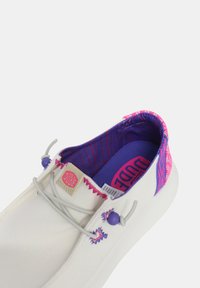 Zapato slip-on de lona blanca con forro de tela en patrones morados y rosas, con cordones elásticos, un cuello texturizado y etiqueta de marca.