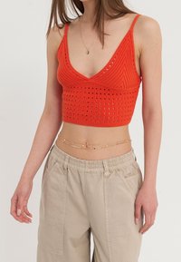 Červený pletený crop top s hlubokým V-výstřihem, zdobený otevřenými pletenými detaily a tenkými ramínky, spárovaný s béžovými kalhotami s elastickým pasem a řetízkovým pásem.