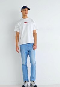 Weißes Kurzarm-T-Shirt mit rotem Levi's-Logo, kombiniert mit hellblauen Jeans und grauen Sneakers. Das Modell trägt eine dunkelblaue Cap.