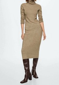 Vestido de punto con cuello alto en beige, ajuste ceñido al cuerpo, mangas largas y un diseño asimétrico. Combinado con botas marrones de caña alta y tacón bloque.