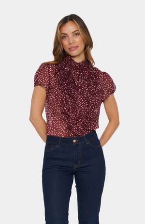Blouse bordeaux transparente avec col à volants et manches courtes, ornée de pois blancs, associée à un jean en denim bleu foncé.