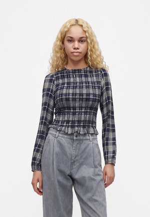 Jeune femme aux longs cheveux blonds bouclés portant un haut ajusté à manches longues bleu marine et blanc à carreaux et un pantalon gris ample sur un fond blanc.