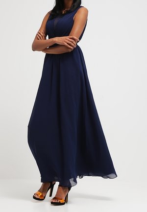 Frau mit verschränkten Armen, die ein ärmelloses, marineblaues Maxikleid und schwarze High Heels mit orangefarbenen Riemen trägt.